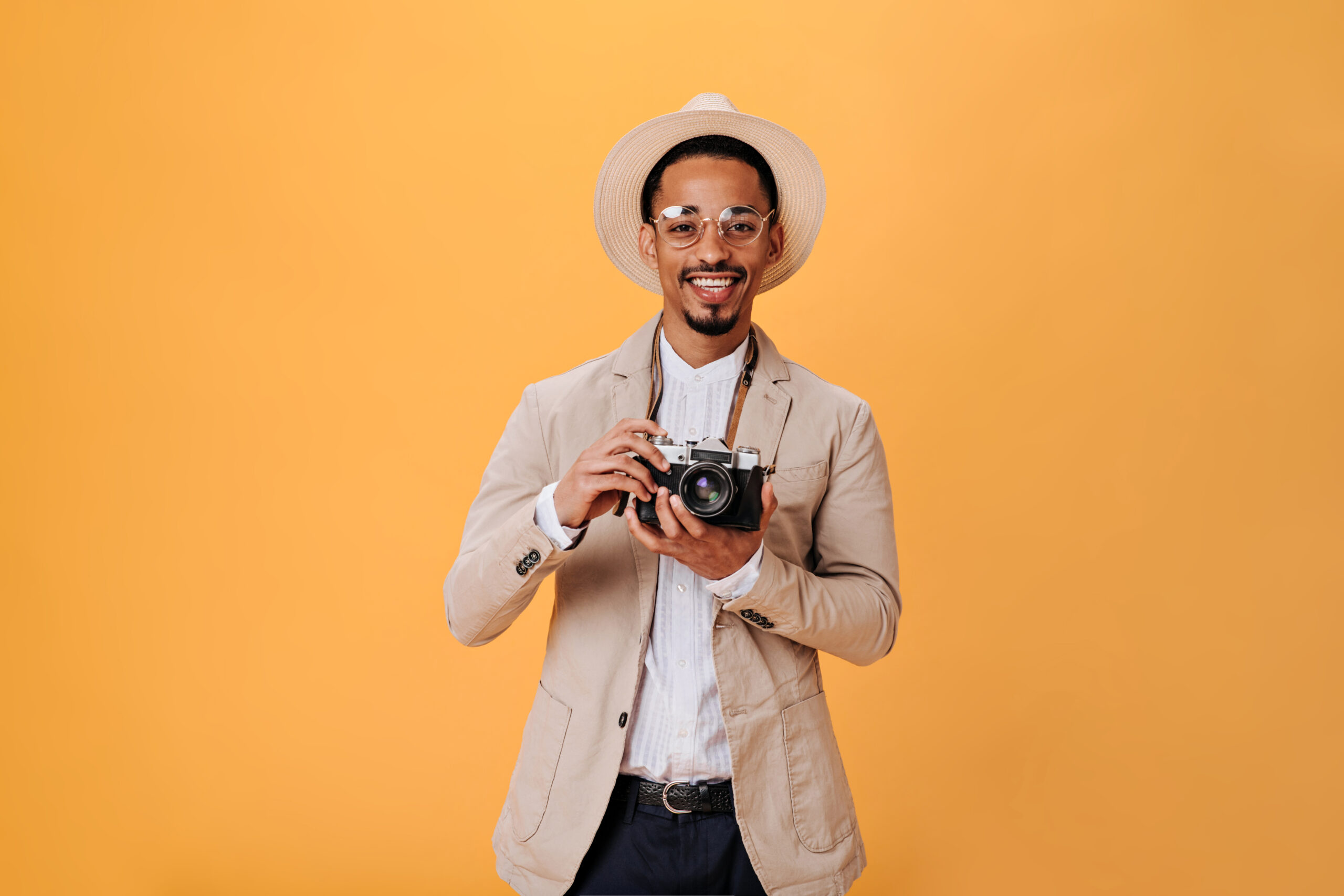 Man in beige hat and jacket makes photo on retro camera. Stylish guy in eyeglasses and white tee smiling on orange background