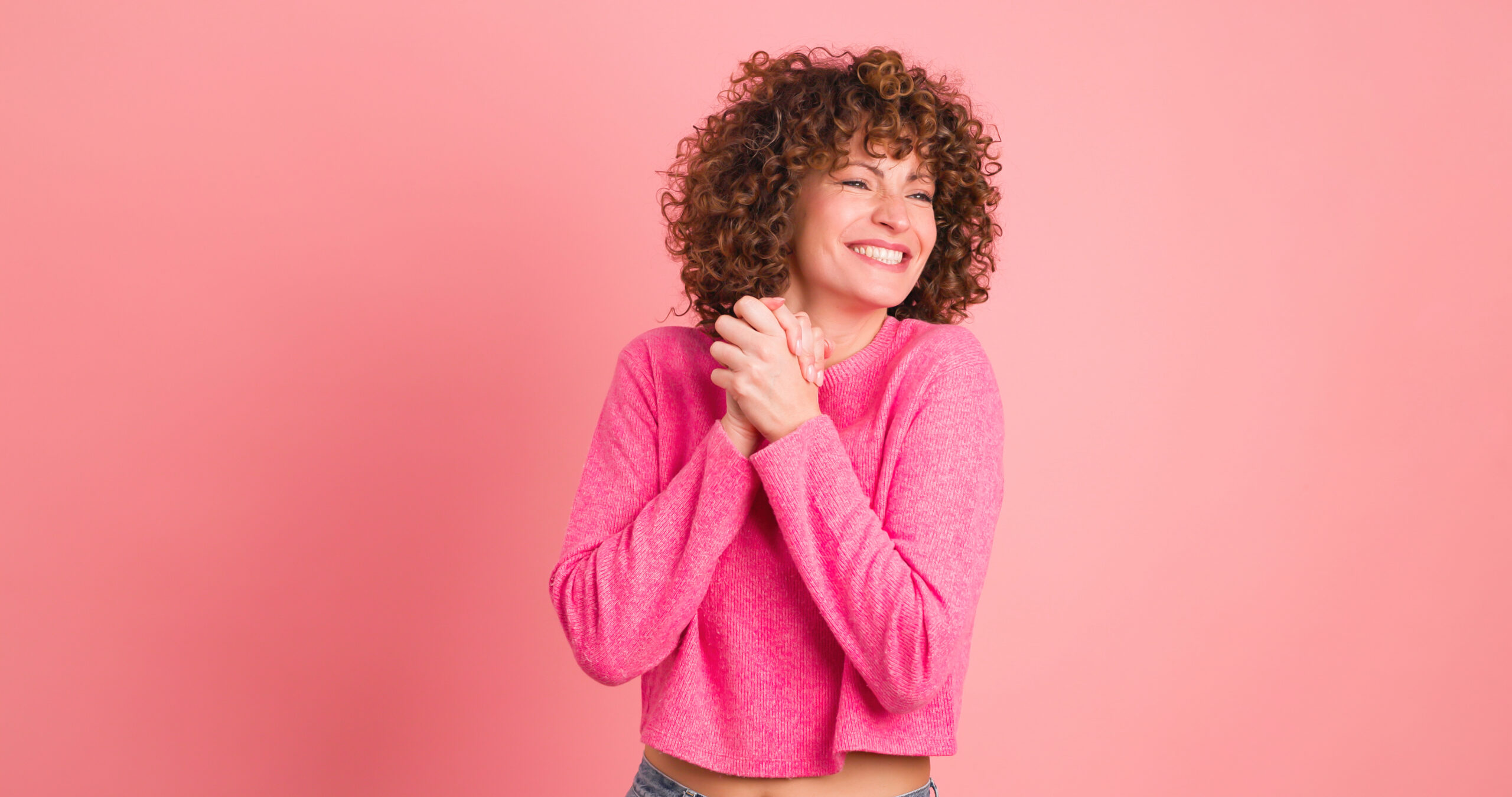 Astonished young curly haired female in makeup and opened mouth looking at camera while standing with folded hands and touching hair on pink background